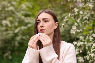 Photo of Woman with paper tissue suffering from seasonal pollen allergy near blossoming shrub on spring day