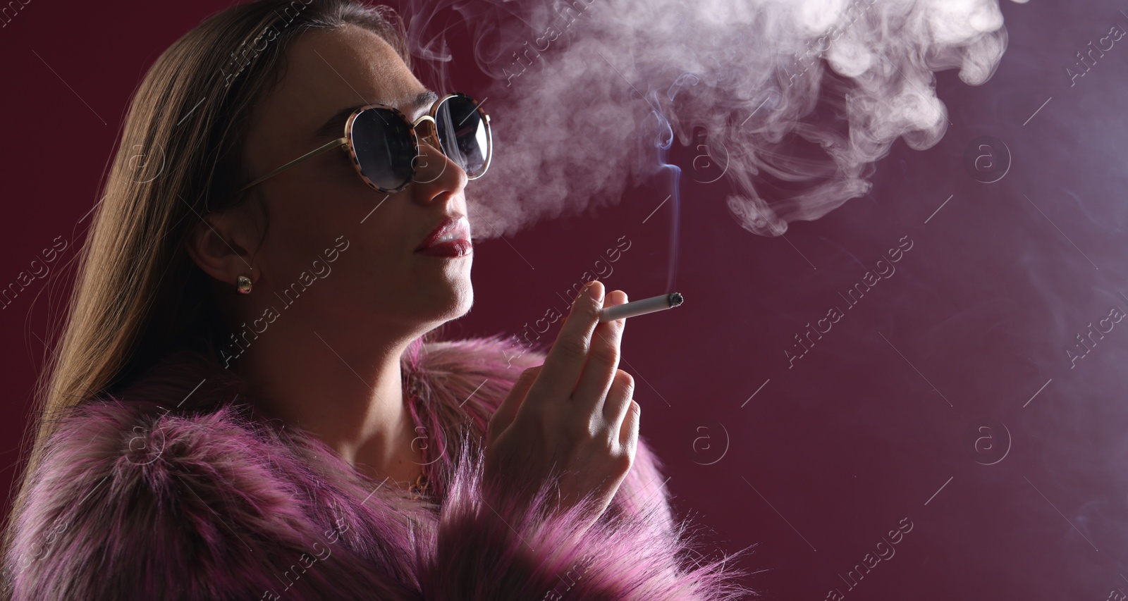 Beautiful woman in fur coat smoking cigarette on dark pink background, closeup Photo of Beautiful woman in fur coat smoking cigarette on dark pink background, closeup
