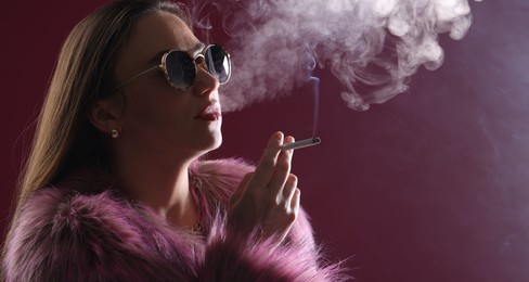 Photo of Beautiful woman in fur coat smoking cigarette on dark pink background, closeup