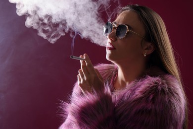 Photo of Beautiful woman in fur coat smoking cigarette on dark pink background, closeup