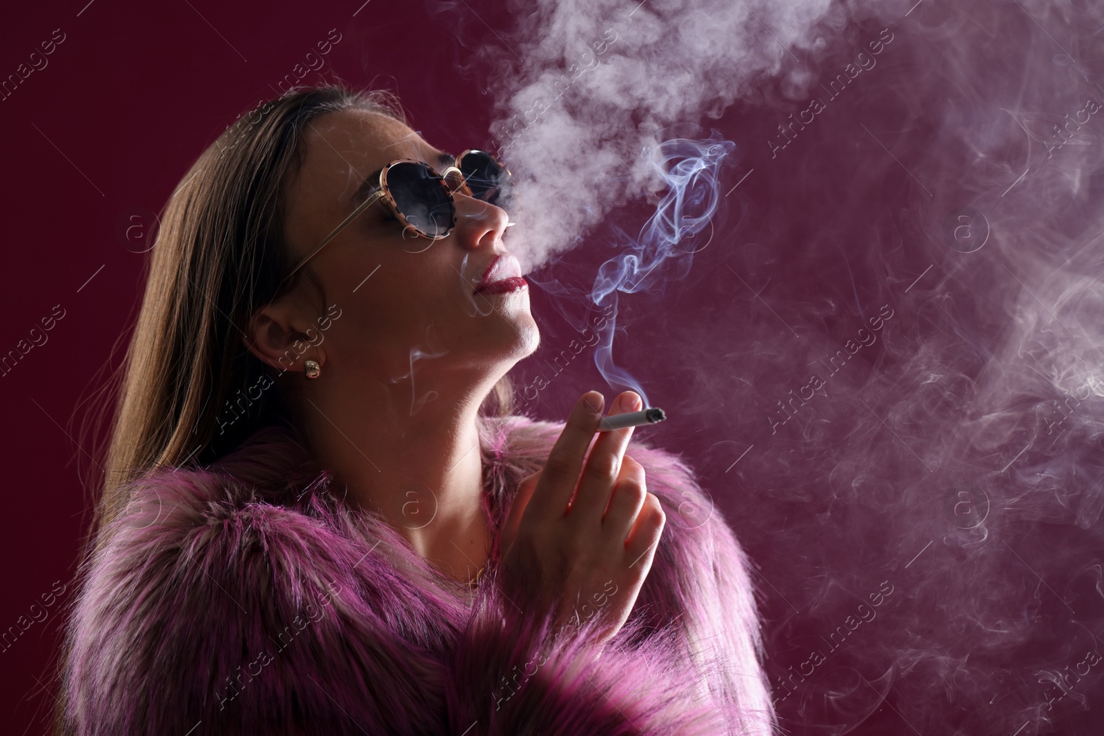 Beautiful woman in fur coat smoking cigarette on dark pink background, closeup Photo of Beautiful woman in fur coat smoking cigarette on dark pink background, closeup