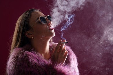 Photo of Beautiful woman in fur coat smoking cigarette on dark pink background, closeup