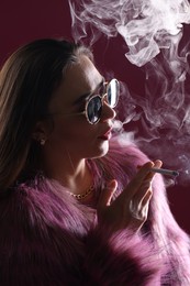 Photo of Beautiful woman in fur coat smoking cigarette on dark pink background, closeup