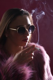 Photo of Beautiful woman in fur coat smoking cigarette on dark pink background, closeup