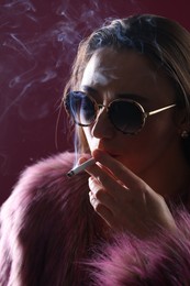 Photo of Beautiful woman in fur coat smoking cigarette on dark pink background, closeup