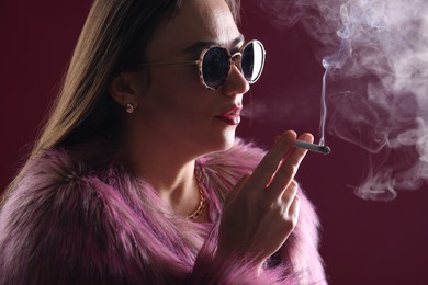 Photo of Beautiful woman in fur coat smoking cigarette on dark pink background, closeup