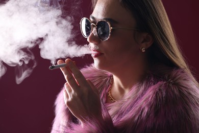 Photo of Beautiful woman in fur coat smoking cigarette on dark pink background, closeup
