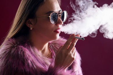 Photo of Beautiful woman in fur coat smoking cigarette on dark pink background, closeup