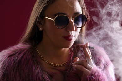 Photo of Beautiful woman in fur coat smoking cigarette on dark pink background, closeup