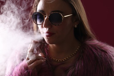 Photo of Beautiful woman in fur coat smoking cigarette on dark pink background, closeup