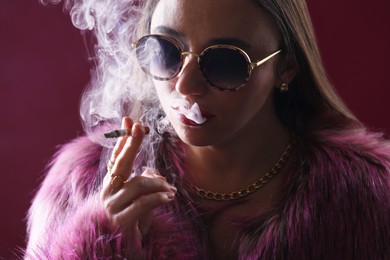 Photo of Beautiful woman in fur coat smoking cigarette on dark pink background, closeup