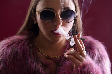 Photo of Beautiful woman in fur coat smoking cigarette on dark pink background, closeup