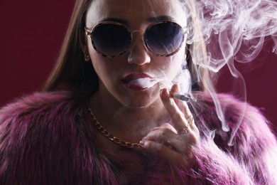 Photo of Beautiful woman in fur coat smoking cigarette on dark pink background, closeup