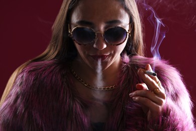 Photo of Beautiful woman in fur coat smoking cigarette on dark pink background, closeup