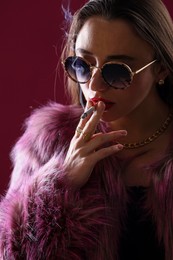 Photo of Beautiful woman in fur coat smoking cigarette on dark pink background, closeup