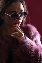 Photo of Beautiful woman in fur coat smoking cigarette on dark pink background, closeup