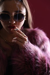 Photo of Beautiful woman in fur coat smoking cigarette on dark pink background, closeup