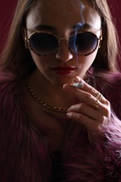 Photo of Beautiful woman in fur coat smoking cigarette on dark pink background, closeup