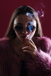 Photo of Beautiful woman in fur coat smoking cigarette on dark pink background, closeup