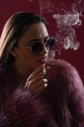 Photo of Beautiful woman in fur coat smoking cigarette on dark pink background, closeup