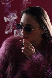 Photo of Beautiful woman in fur coat smoking cigarette on dark pink background, closeup