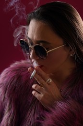 Photo of Beautiful woman in fur coat smoking cigarette on dark pink background, closeup