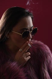 Photo of Beautiful woman in fur coat smoking cigarette on dark pink background, closeup