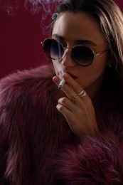 Photo of Beautiful woman in fur coat smoking cigarette on dark pink background, closeup