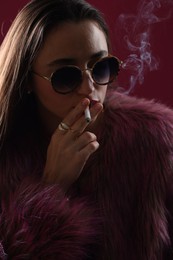 Photo of Beautiful woman in fur coat smoking cigarette on dark pink background, closeup