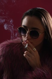 Photo of Beautiful woman in fur coat smoking cigarette on dark pink background, closeup