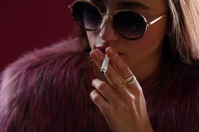 Photo of Beautiful woman in fur coat smoking cigarette on dark pink background, closeup