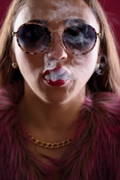 Photo of Beautiful woman in fur coat smoking cigarette on dark pink background, closeup