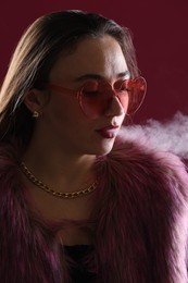 Photo of Beautiful woman in fur coat smoking cigarette on dark pink background, closeup