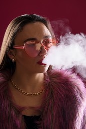 Photo of Beautiful woman in fur coat smoking cigarette on dark pink background, closeup