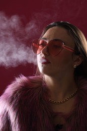 Photo of Beautiful woman in fur coat smoking cigarette on dark pink background, closeup