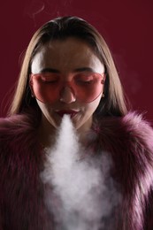 Photo of Beautiful woman in fur coat smoking cigarette on dark pink background, closeup