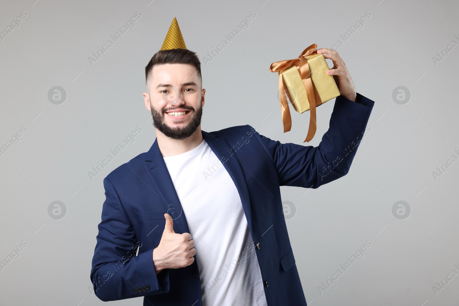Happy Birthday. Handsome man with party hat and gift box showing thumbs up on grey background Photo of Happy Birthday. Handsome man with party hat and gift box showing thumbs up on grey background