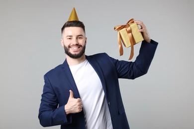 Photo of Happy Birthday. Handsome man with party hat and gift box showing thumbs up on grey background