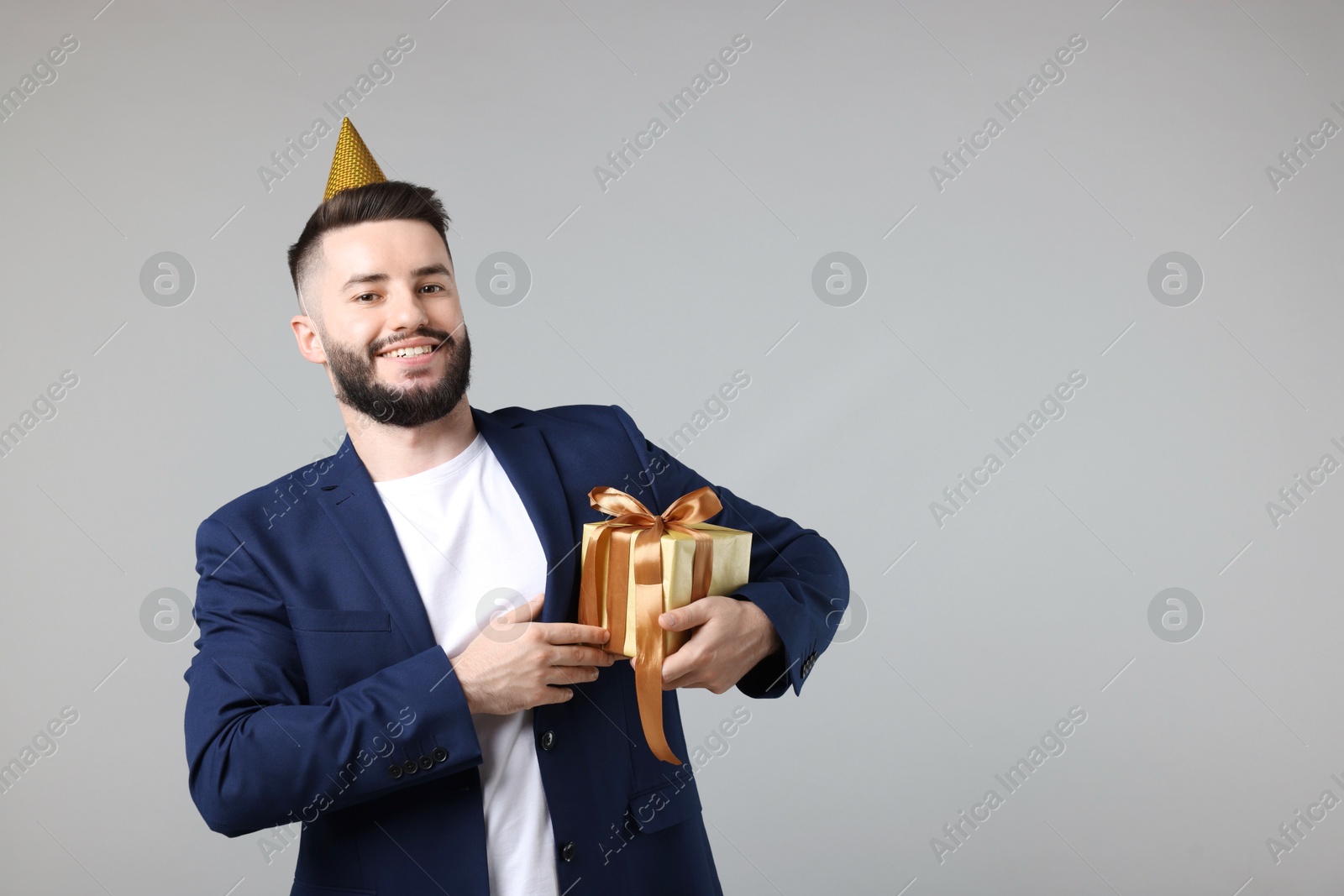 Happy Birthday. Handsome man with party hat and gift box on grey background, space for text Photo of Happy Birthday. Handsome man with party hat and gift box on grey background, space for text