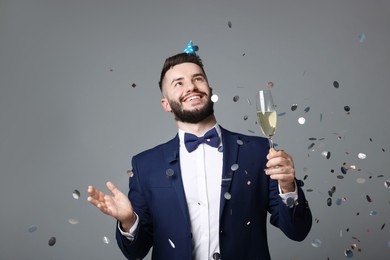 Photo of Happy Birthday. Handsome man with party hat and glass of champagne under confetti shower on grey background