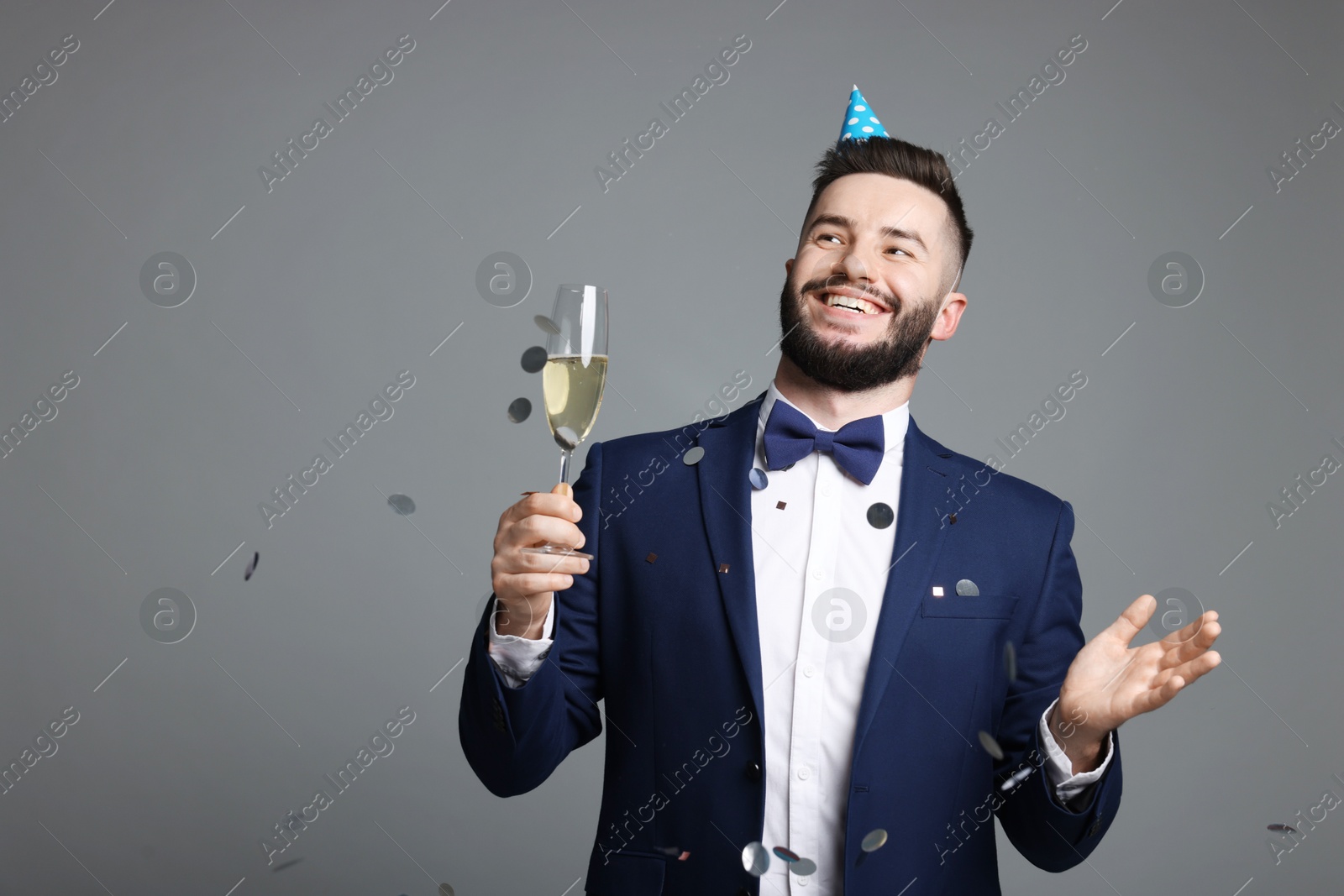 Happy Birthday. Handsome man with party hat and glass of champagne under confetti shower on grey background, space for text Photo of Happy Birthday. Handsome man with party hat and glass of champagne under confetti shower on grey background, space for text