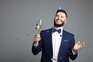 Photo of Happy Birthday. Handsome man with party hat and glass of champagne under confetti shower on grey background, space for text