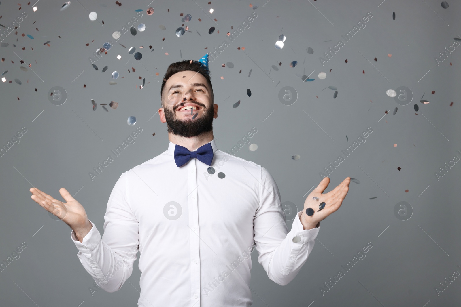 Happy Birthday. Handsome man with party hat under confetti shower on grey background, space for text Photo of Happy Birthday. Handsome man with party hat under confetti shower on grey background, space for text
