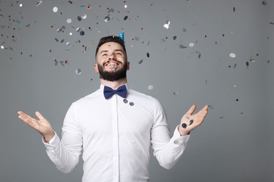 Photo of Happy Birthday. Handsome man with party hat under confetti shower on grey background, space for text