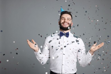 Photo of Happy Birthday. Handsome man with party hat under confetti shower on grey background, space for text