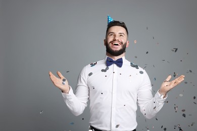 Photo of Happy Birthday. Handsome man with party hat under confetti shower on grey background, space for text