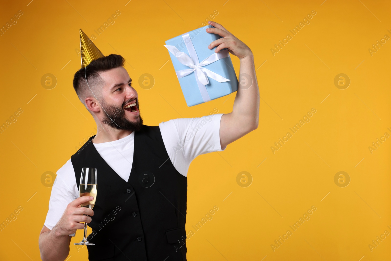 Happy Birthday. Handsome man with party hat, glass of champagne and gift box on orange background, space for text Photo of Happy Birthday. Handsome man with party hat, glass of champagne and gift box on orange background, space for text
