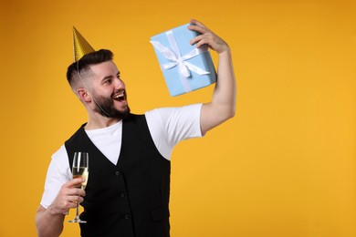 Photo of Happy Birthday. Handsome man with party hat, glass of champagne and gift box on orange background, space for text