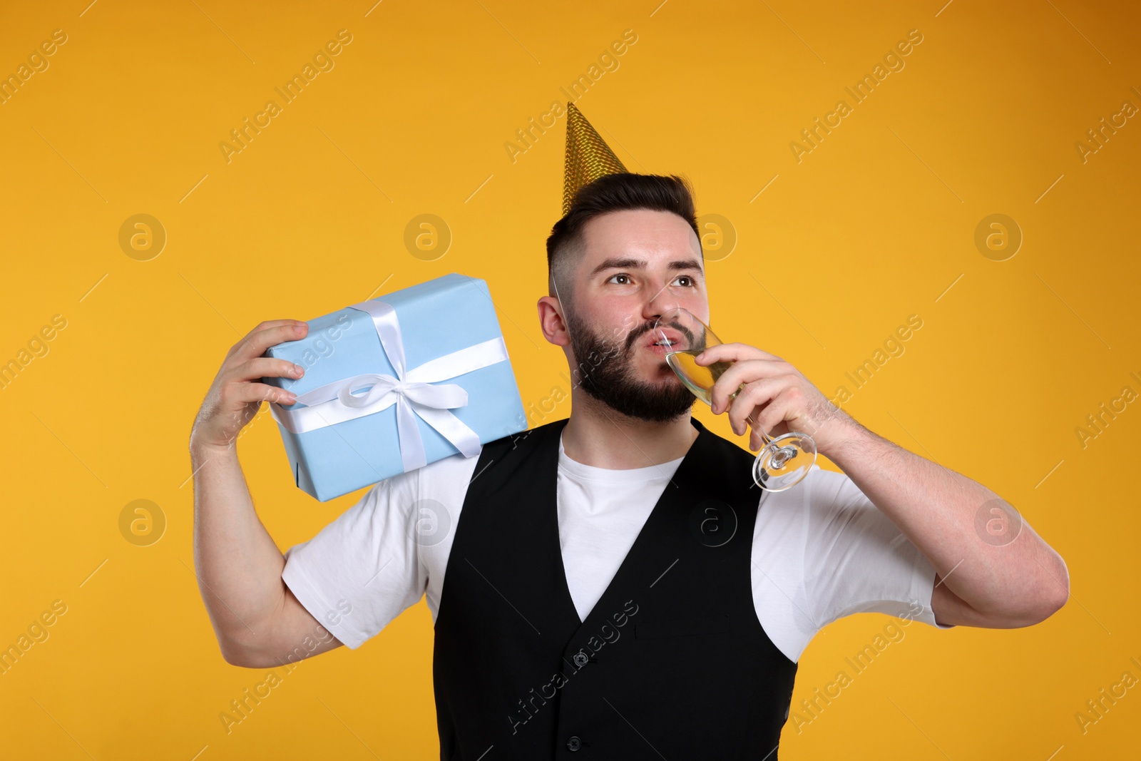 Happy Birthday. Handsome man with party hat, glass of champagne and gift box on orange background Photo of Happy Birthday. Handsome man with party hat, glass of champagne and gift box on orange background