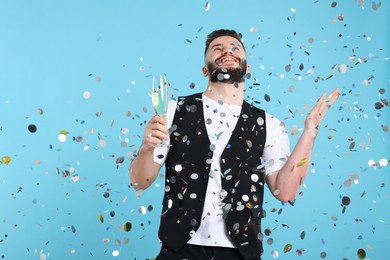 Photo of Happy Birthday. Handsome man with glass of champagne under confetti shower on light blue background, space for text